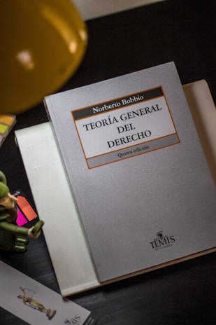A book titled 'Teoría General del Derecho' by Norberto Bobbio is placed on a dark surface next to a desk lamp. The book appears to be a fifth edition from Editor Temis. A small toy soldier, a colorful bookmark, and a folded white notebook are also visible in the scene.