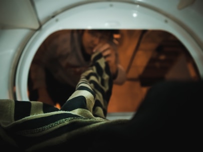 A person is reaching into a washing machine drum, grabbing a striped garment. The view is from inside the machine looking out. The lighting is dim, creating a warm, enclosed atmosphere.