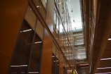 A modern interior featuring high ceilings and rows of glass-panelled walls, creating a spacious, well-lit corridor. The architectural design incorporates clean lines, with balconies and lighting fixtures enhancing the sense of openness.