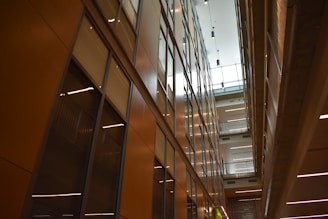 A modern interior featuring high ceilings and rows of glass-panelled walls, creating a spacious, well-lit corridor. The architectural design incorporates clean lines, with balconies and lighting fixtures enhancing the sense of openness.