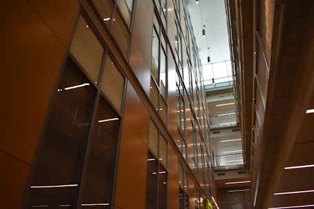 A modern interior featuring high ceilings and rows of glass-panelled walls, creating a spacious, well-lit corridor. The architectural design incorporates clean lines, with balconies and lighting fixtures enhancing the sense of openness.
