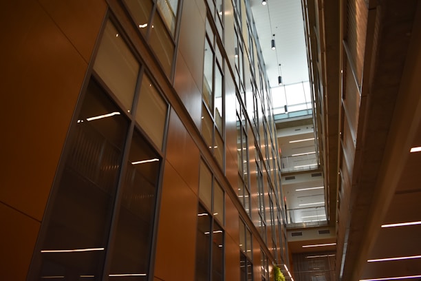 A modern interior featuring high ceilings and rows of glass-panelled walls, creating a spacious, well-lit corridor. The architectural design incorporates clean lines, with balconies and lighting fixtures enhancing the sense of openness.