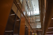 A modern interior featuring high ceilings and rows of glass-panelled walls, creating a spacious, well-lit corridor. The architectural design incorporates clean lines, with balconies and lighting fixtures enhancing the sense of openness.