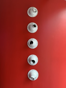 Five white circular fixtures are aligned vertically on a bright red surface. Each fixture resembles a mounting bracket or a vent outlet, with circular openings.