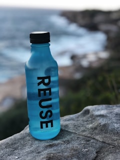 A blue bottle with the word 'REUSE' vertically printed on it is placed on a rock. In the background, there is a scenic view of the ocean with waves crashing against the rocky shore and a blurred coastline.