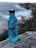 A blue bottle with the word 'REUSE' vertically printed on it is placed on a rock. In the background, there is a scenic view of the ocean with waves crashing against the rocky shore and a blurred coastline.