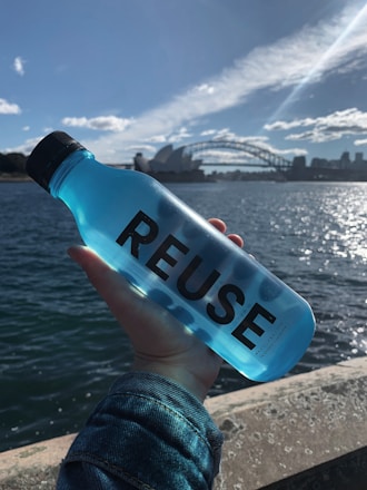 A hand holds a translucent blue bottle with the word 'REUSE' printed on it, against a backdrop of a large body of water and a city skyline. Notable landmarks such as a famous opera house and bridge are visible under a clear blue sky with scattered clouds.