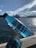 A hand holds a translucent blue bottle with the word 'REUSE' printed on it, against a backdrop of a large body of water and a city skyline. Notable landmarks such as a famous opera house and bridge are visible under a clear blue sky with scattered clouds.