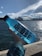 A hand holds a translucent blue bottle with the word 'REUSE' printed on it, against a backdrop of a large body of water and a city skyline. Notable landmarks such as a famous opera house and bridge are visible under a clear blue sky with scattered clouds.