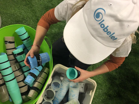 A man is sorting reusable plastic cups into two different bins on a grassy surface, wearing a white cap with a blue logo.