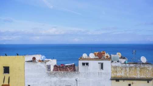 Several buildings with weathered paint and numerous satellite dishes on their roofs overlook a vast, calm sea under a clear blue sky. A distant horizon separates water from sky.