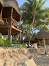 A tropical beach setting with a wooden thatch-roofed structure elevated on stilts, surrounded by lush green palm trees. The sandy beach is dotted with resort-style thatched umbrellas and lounge chairs.