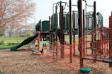 A playground structure made of dark green and brown materials, featuring slides, climbing bars, and platforms. The area is surrounded by bright orange safety fencing, indicating construction or maintenance work. The ground is covered with wood chips, and nearby, there is a grassy field and trees with budding leaves.