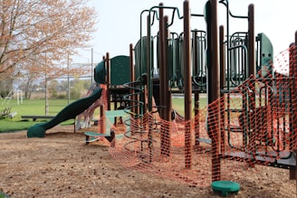 A playground structure made of dark green and brown materials, featuring slides, climbing bars, and platforms. The area is surrounded by bright orange safety fencing, indicating construction or maintenance work. The ground is covered with wood chips, and nearby, there is a grassy field and trees with budding leaves.