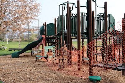 A playground structure made of dark green and brown materials, featuring slides, climbing bars, and platforms. The area is surrounded by bright orange safety fencing, indicating construction or maintenance work. The ground is covered with wood chips, and nearby, there is a grassy field and trees with budding leaves.