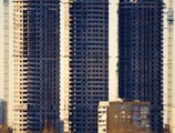 Three tall, unfinished skyscrapers stand closely together, displaying their raw concrete structure and numerous identical windows. In front of them, a smaller residential building with a similar window pattern is visible. Several cranes are positioned around the buildings, suggesting ongoing construction. Shadows and sunlight highlight the edges of the structures.