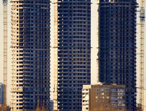 Three tall, unfinished skyscrapers stand closely together, displaying their raw concrete structure and numerous identical windows. In front of them, a smaller residential building with a similar window pattern is visible. Several cranes are positioned around the buildings, suggesting ongoing construction. Shadows and sunlight highlight the edges of the structures.