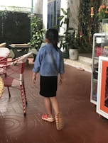 A child wearing a denim jacket and a black dress is walking on a brown tiled floor. The child is barefoot on one foot and wearing an orange shoe on the other. The setting appears to be a patio or garden with several plants in ceramic pots. There are wicker chairs with red and white patterns on the left and an object on the right, possibly a cabinet.