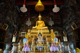 A richly decorated temple interior with a large golden Buddha statue at the center, flanked by smaller statues. The walls are adorned with intricate murals, and crystal chandeliers hang from the ceiling. Various ornate offerings and ceremonial items are placed on tables in front of the statues.