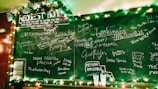 A chalkboard filled with handwritten movie titles and requests. The top is adorned with Christmas lights and garland, creating a festive atmosphere. Two empty plastic cups sit on a ledge beneath the board.