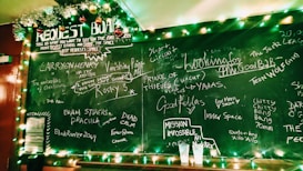 A chalkboard filled with handwritten movie titles and requests. The top is adorned with Christmas lights and garland, creating a festive atmosphere. Two empty plastic cups sit on a ledge beneath the board.