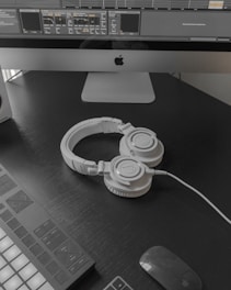 A modern workspace setup featuring a large computer monitor displaying music production software. Below the screen, a pair of white over-ear headphones lies on a dark desk surface. To the left, part of a MIDI controller is visible. A wireless mouse rests nearby.