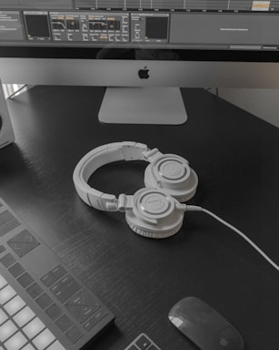A modern workspace setup featuring a large computer monitor displaying music production software. Below the screen, a pair of white over-ear headphones lies on a dark desk surface. To the left, part of a MIDI controller is visible. A wireless mouse rests nearby.