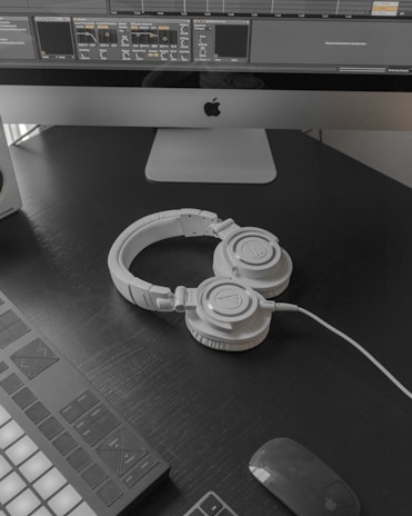 A modern workspace setup featuring a large computer monitor displaying music production software. Below the screen, a pair of white over-ear headphones lies on a dark desk surface. To the left, part of a MIDI controller is visible. A wireless mouse rests nearby.