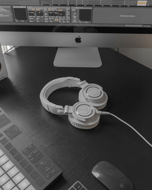 A modern workspace setup featuring a large computer monitor displaying music production software. Below the screen, a pair of white over-ear headphones lies on a dark desk surface. To the left, part of a MIDI controller is visible. A wireless mouse rests nearby.