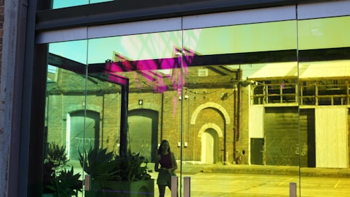A large glass window reflects an urban street scene, with yellow and pink hues tinting the reflection. The window showcases brick buildings with arched doorways and large garage doors. Plants are visible inside the window, and a person is walking outside, partially obscured by the glass.