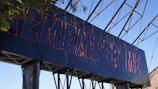 A large structure displays the words 'REMEMBER ME' in bold, red-outlined letters against a blue background. The structure is set outdoors with visible metal framework and some foliage partially covering the corner.