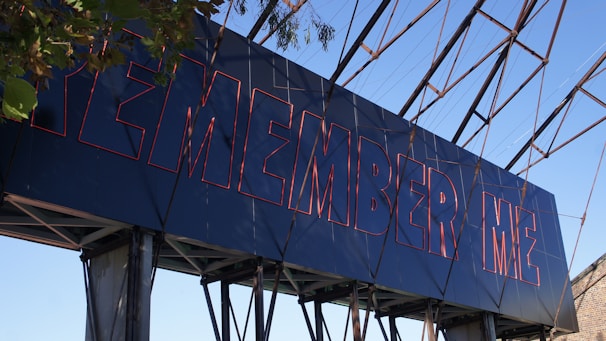 A large structure displays the words 'REMEMBER ME' in bold, red-outlined letters against a blue background. The structure is set outdoors with visible metal framework and some foliage partially covering the corner.