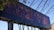 A large structure displays the words 'REMEMBER ME' in bold, red-outlined letters against a blue background. The structure is set outdoors with visible metal framework and some foliage partially covering the corner.