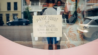 A handwritten sign taped to a glass window, requesting people not to sit on the windowsill as repairs are needed. Behind the glass, several people and vehicles on the street are visible.