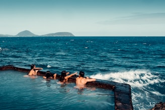 Several people are relaxing in a natural pool overlooking the ocean. The pool is situated on a rocky outcrop, and in the background, the vast sea stretches towards distant islands. The scene is tranquil and serene, capturing the essence of a seaside retreat.