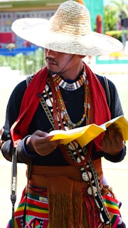 A person wearing a large straw hat, ornate clothing with colorful patterns, beaded necklaces, and a red scarf reads a yellow pamphlet. A camera strap is visible over the shoulder, suggesting a photography interest. The background is blurred, indicating an outdoor setting with vibrant colors.