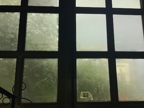 A large grid of frosted glass windows with a dark frame. The glass is slightly foggy or dirty, allowing limited visibility through it. Soft, diffused light filters in through the windows, creating a muted atmosphere. A black metal railing with a decorative swirl is partially visible in the lower left corner.