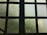 A large grid of frosted glass windows with a dark frame. The glass is slightly foggy or dirty, allowing limited visibility through it. Soft, diffused light filters in through the windows, creating a muted atmosphere. A black metal railing with a decorative swirl is partially visible in the lower left corner.