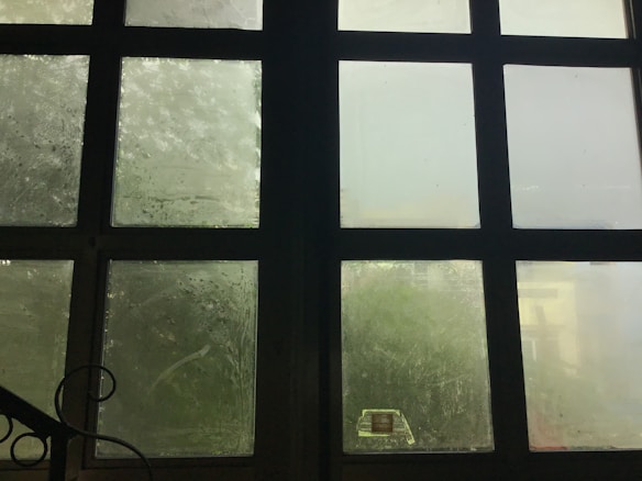 A large grid of frosted glass windows with a dark frame. The glass is slightly foggy or dirty, allowing limited visibility through it. Soft, diffused light filters in through the windows, creating a muted atmosphere. A black metal railing with a decorative swirl is partially visible in the lower left corner.