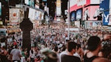 A bustling urban scene filled with a large crowd of people in a public square, surrounded by bright and colorful electronic billboards displaying advertisements. The area is lively and packed with tourists and locals engaging in various activities.