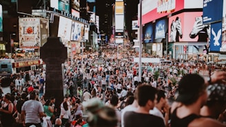 A bustling urban scene filled with a large crowd of people in a public square, surrounded by bright and colorful electronic billboards displaying advertisements. The area is lively and packed with tourists and locals engaging in various activities.