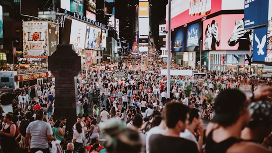 A bustling urban scene filled with a large crowd of people in a public square, surrounded by bright and colorful electronic billboards displaying advertisements. The area is lively and packed with tourists and locals engaging in various activities.
