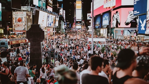 A bustling urban scene filled with a large crowd of people in a public square, surrounded by bright and colorful electronic billboards displaying advertisements. The area is lively and packed with tourists and locals engaging in various activities.