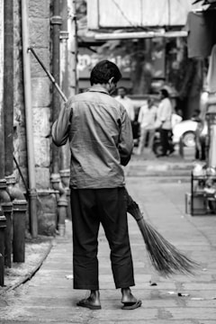 A person is sweeping a street in an urban setting. The individual is dressed in casual attire and is holding a broom made of natural materials. The background features some buildings and a few people blurred out in motion, suggesting a busy street atmosphere.