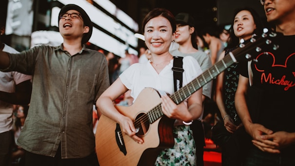 A group of people is gathered in what appears to be a lively, outdoor setting. A woman in the foreground is smiling while holding a guitar. Others around her are engaged, some looking in different directions, suggesting movement and interaction among them. The atmosphere seems vibrant and joyful, possibly indicating a musical or festive event.