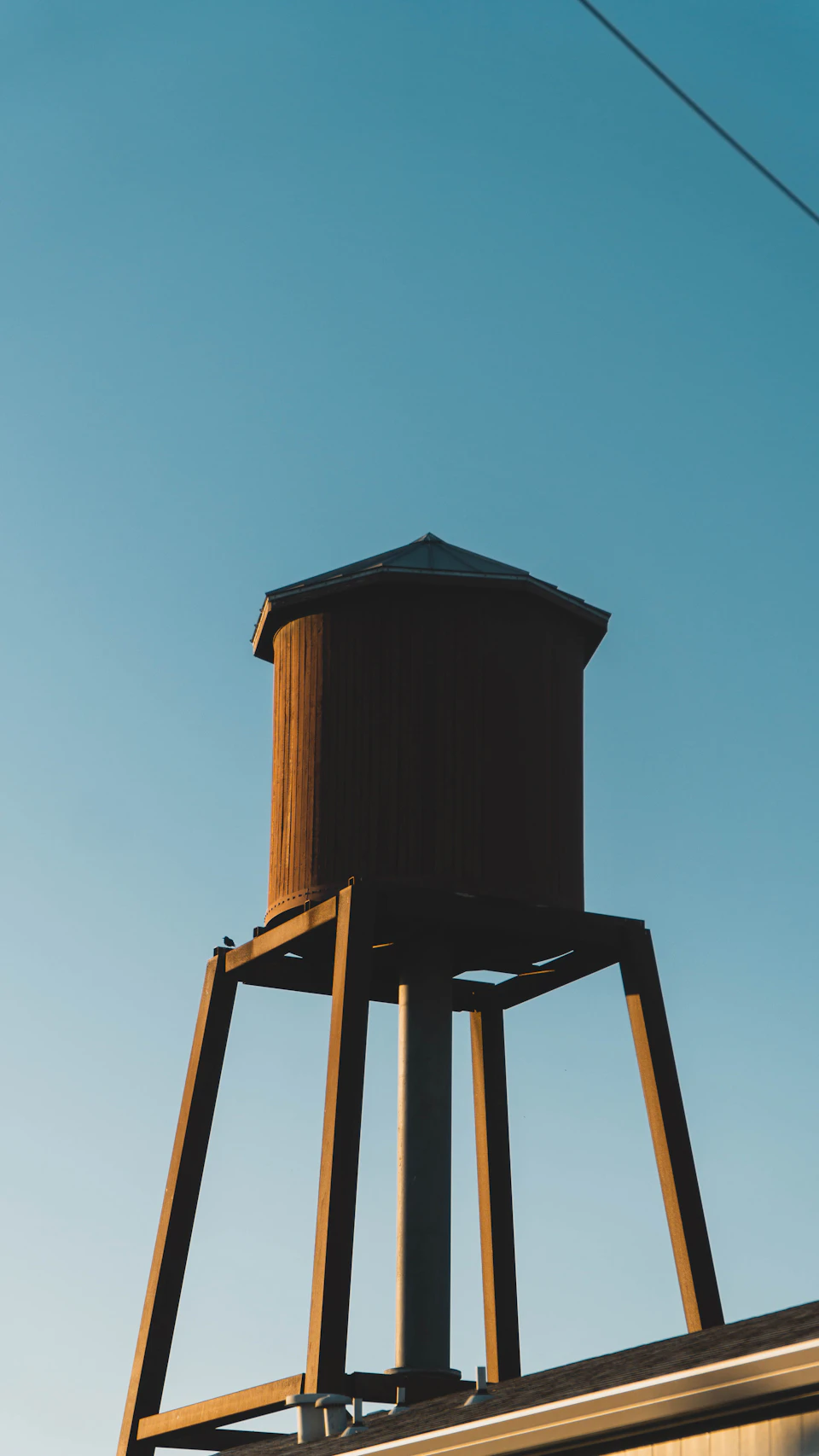 The image depicts a large, brown water tower standing tall against a clear blue sky. The water tower is situated on a wooden platform with four legs and a triangular roof, giving it an imposing presence. The perspective of the photo is from below, looking up at the water tower, which enhances its grandeur. In the background, there are power lines running across the scene, adding to the industrial feel of the image.