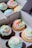 A close-up view of a box containing six colorful cupcakes. Each cupcake features swirls of pastel-colored frosting in pink, green, blue, and white. Some cupcakes are decorated with carrot-shaped fondant pieces and other small, festive decorations. The cupcakes come in both chocolate and vanilla flavors and are arranged neatly in a white cardboard box with dividers.