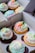 A close-up view of a box containing six colorful cupcakes. Each cupcake features swirls of pastel-colored frosting in pink, green, blue, and white. Some cupcakes are decorated with carrot-shaped fondant pieces and other small, festive decorations. The cupcakes come in both chocolate and vanilla flavors and are arranged neatly in a white cardboard box with dividers.