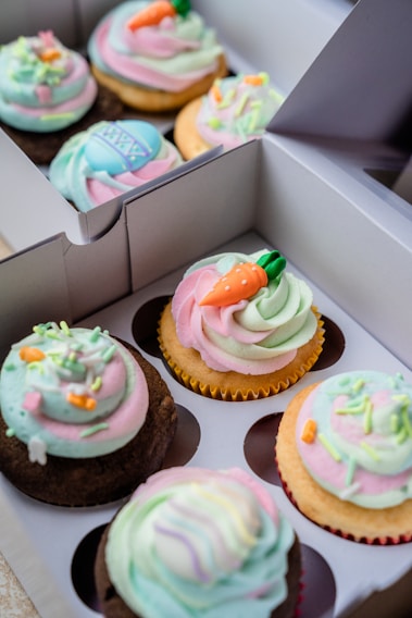 A close-up view of a box containing six colorful cupcakes. Each cupcake features swirls of pastel-colored frosting in pink, green, blue, and white. Some cupcakes are decorated with carrot-shaped fondant pieces and other small, festive decorations. The cupcakes come in both chocolate and vanilla flavors and are arranged neatly in a white cardboard box with dividers.