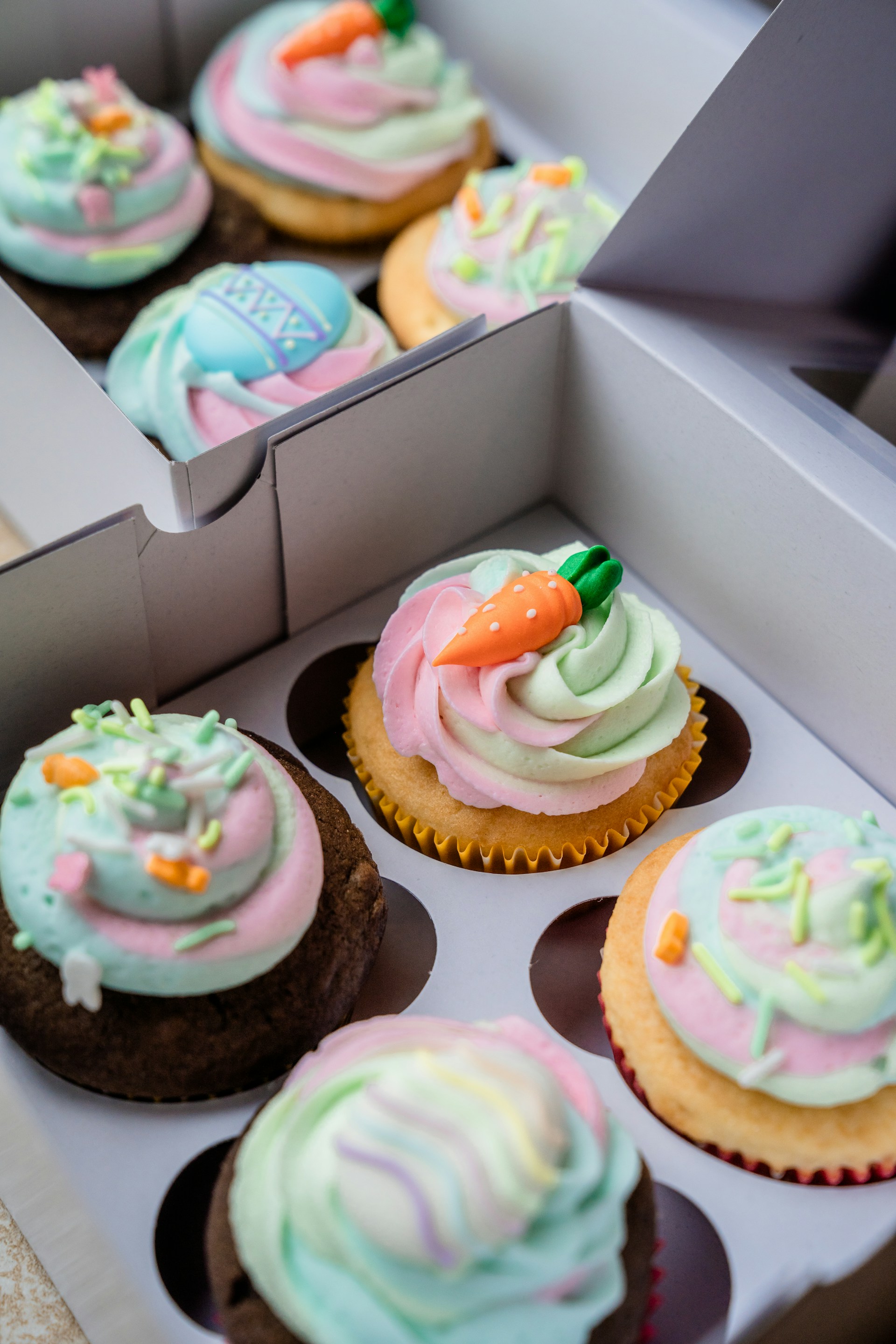 A close-up view of a box containing six colorful cupcakes. Each cupcake features swirls of pastel-colored frosting in pink, green, blue, and white. Some cupcakes are decorated with carrot-shaped fondant pieces and other small, festive decorations. The cupcakes come in both chocolate and vanilla flavors and are arranged neatly in a white cardboard box with dividers.